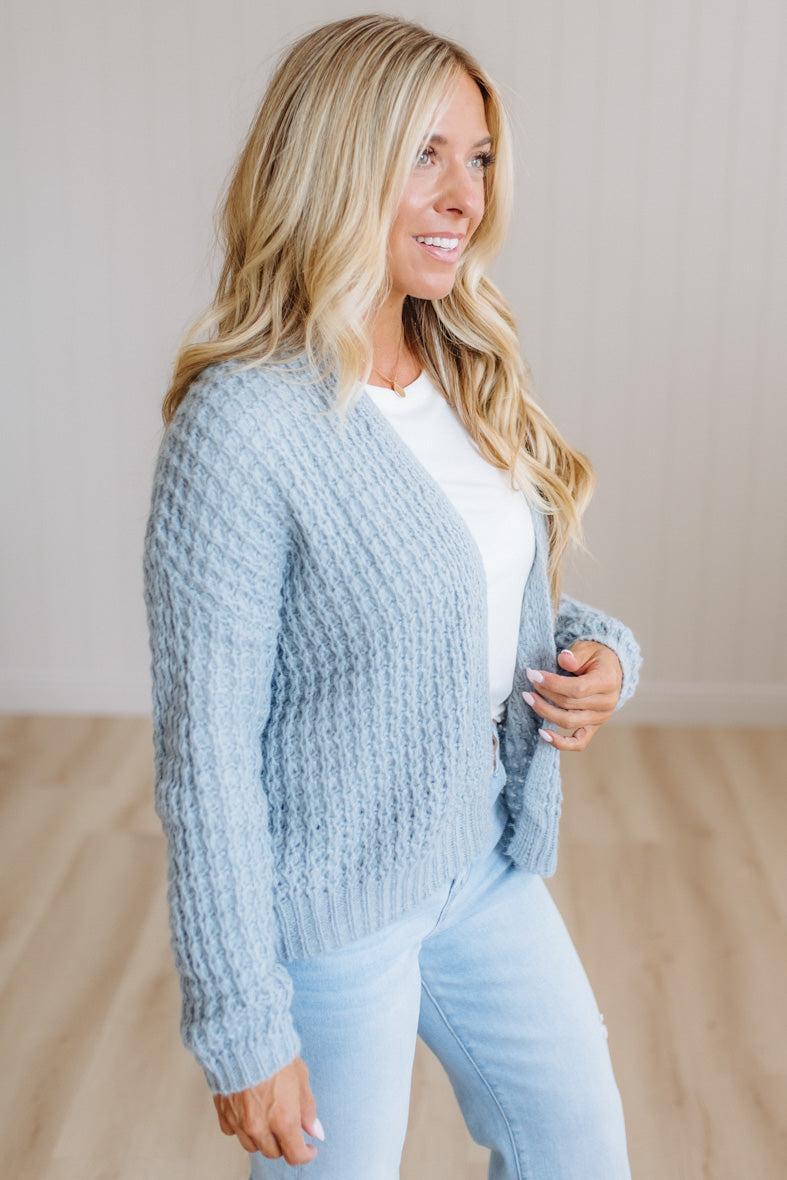 A woman wearing a light blue chunky cable-knit cardigan over a white top and light blue jeans, standing in a minimalist room with light wood floors and a beige wall.