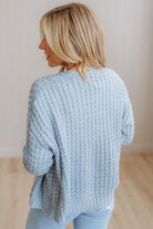 A woman with blonde hair is shown from behind wearing a light blue chunky cable-knit sweater and light blue jeans in a brightly lit indoor setting.