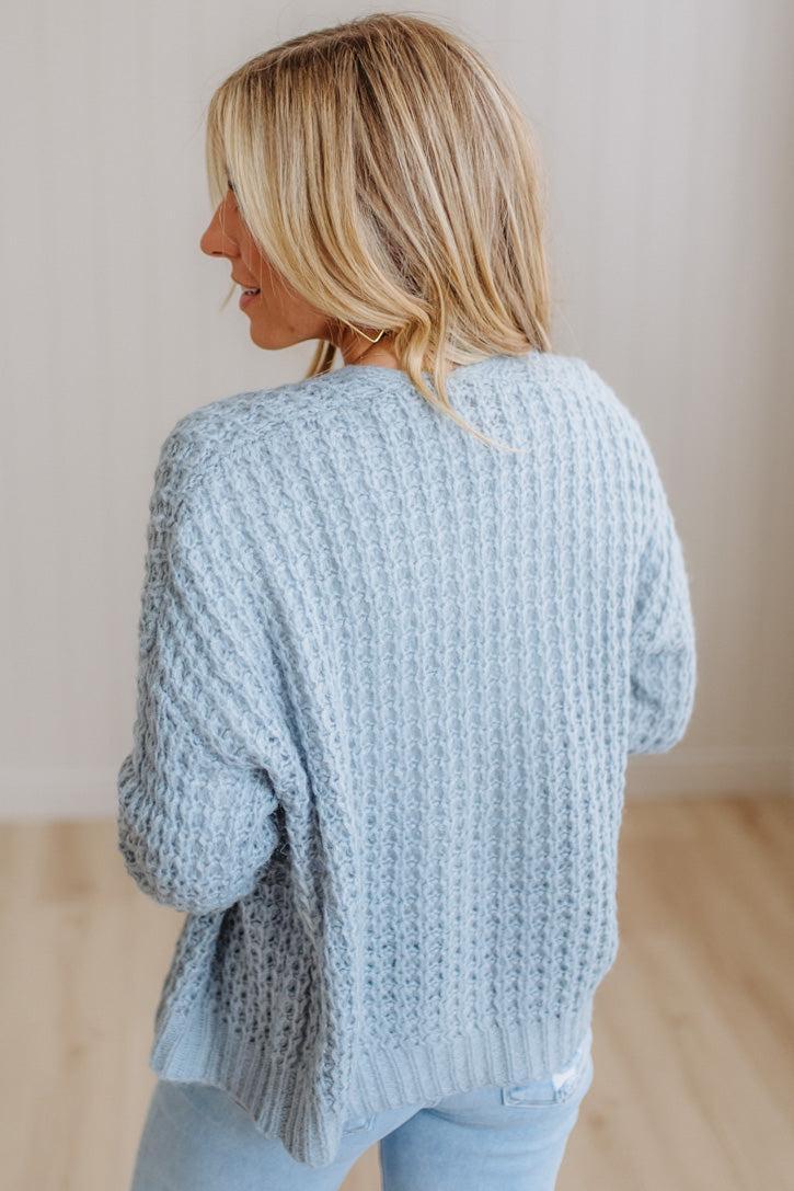 A woman with blonde hair is shown from behind wearing a light blue chunky cable-knit sweater and light blue jeans in a brightly lit indoor setting.