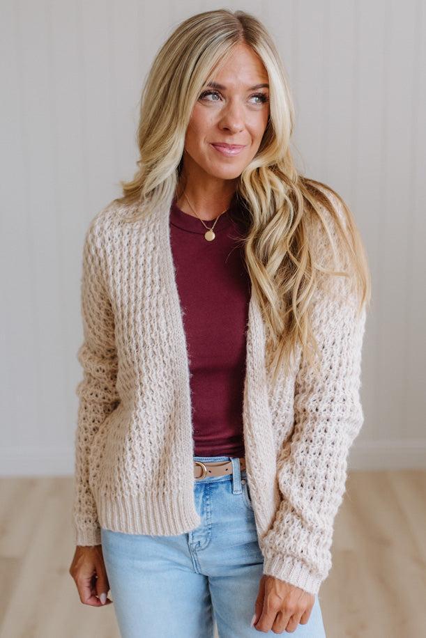 A woman with long blonde hair wearing a cream cable-knit cardigan over a burgundy top and light blue jeans, standing against a light-colored paneled wall.