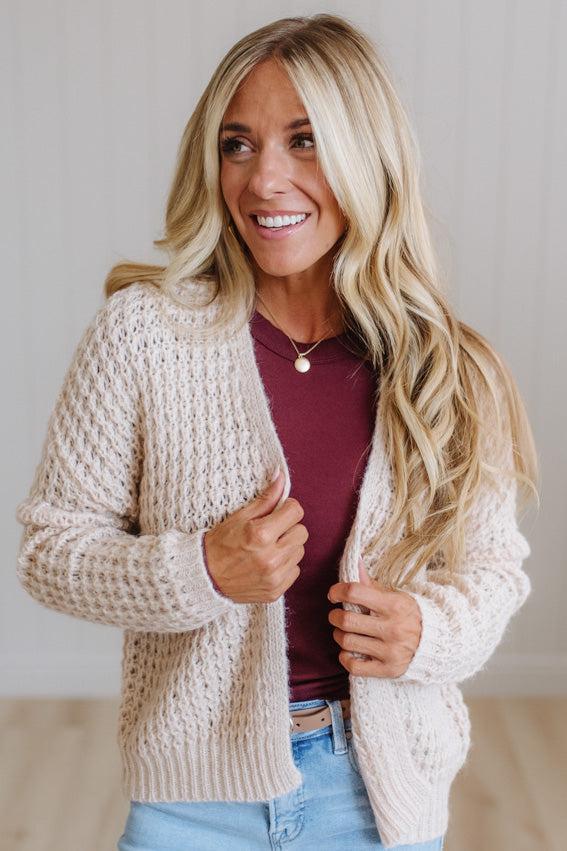 A smiling blonde woman wearing a chunky cream cable-knit cardigan over a burgundy top and light blue jeans, standing against a light neutral wall.