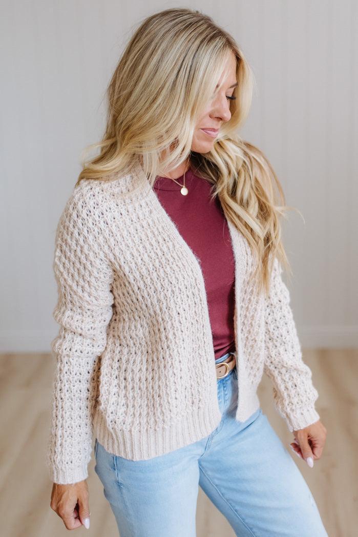 A woman with long blonde hair wearing a cream cable-knit cardigan over a burgundy top and light blue jeans, standing against a light-colored paneled wall.
