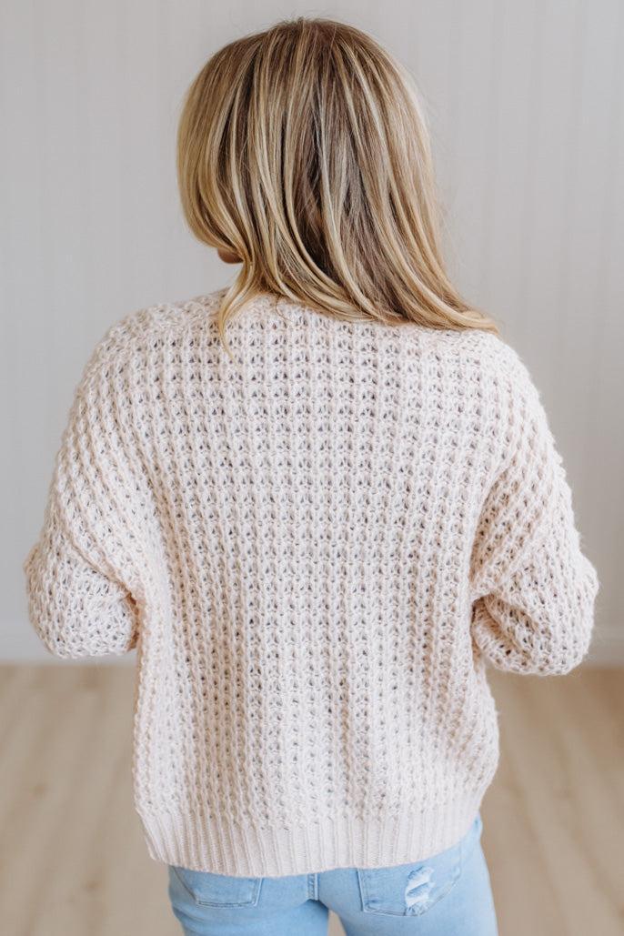 A woman with long blonde hair wearing a cream cable-knit cardigan over a burgundy top and light blue jeans, standing against a light-colored paneled wall.