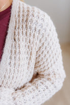Close-up of a person wearing a chunky knit cardigan in a warm off-white or cream color, layered over a burgundy top. The cardigan features a textured open-knit pattern and is shown against a softly blurred neutral background.