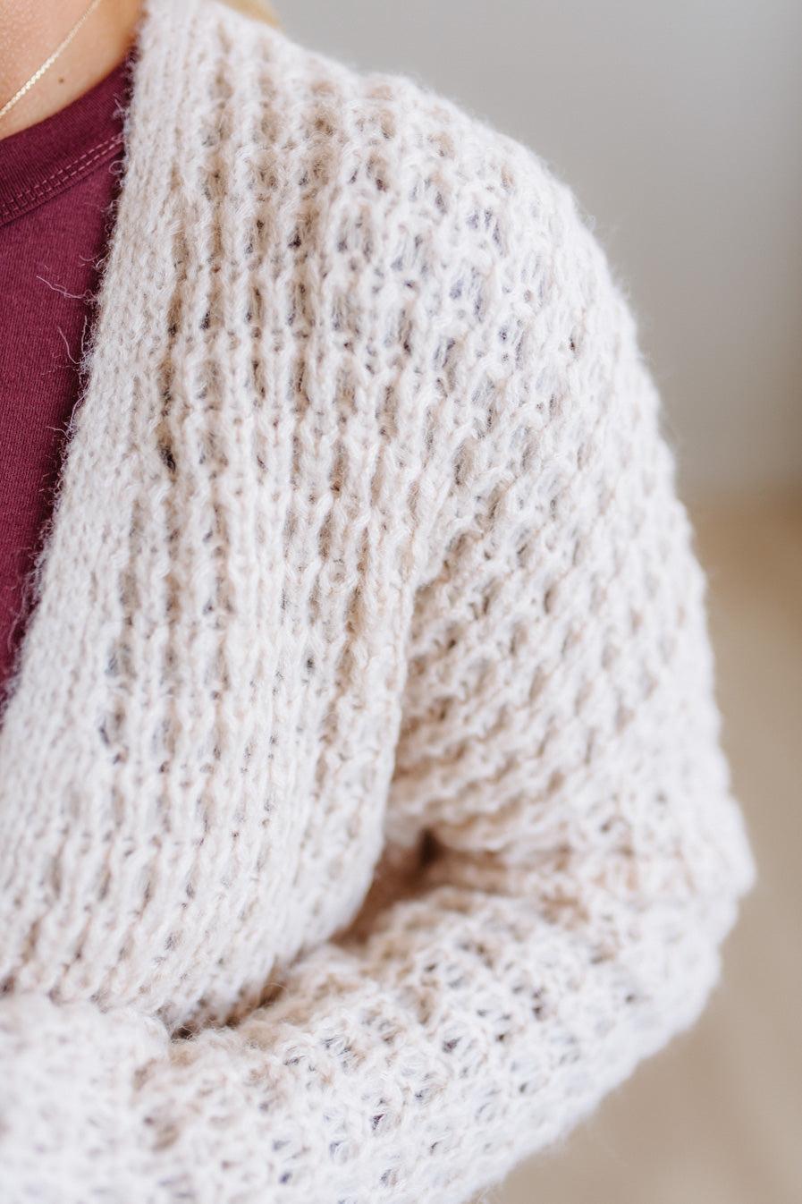 Close-up of a person wearing a chunky knit cardigan in a warm off-white or cream color, layered over a burgundy top. The cardigan features a textured open-knit pattern and is shown against a softly blurred neutral background.