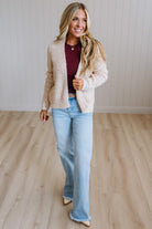 A woman modeling a cream cable-knit cardigan over a burgundy top and light-wash flare jeans, standing on a light wood floor against a white paneled wall.