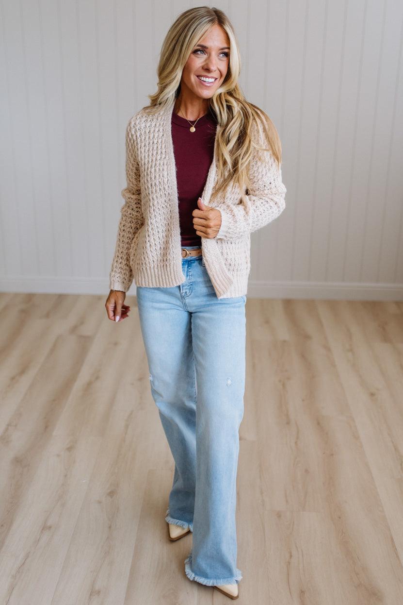 A woman modeling a cream cable-knit cardigan over a burgundy top and light-wash flare jeans, standing on a light wood floor against a white paneled wall.