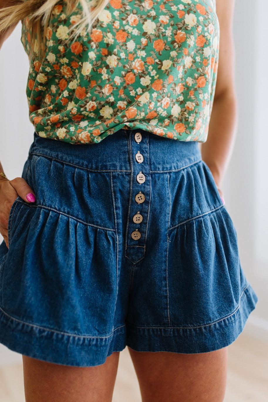 Woman in a green floral tank wearing dark wash denim shorts with wooden buttons at the front, and tiered flare cut.