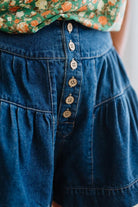 Woman in a green floral tank wearing dark wash denim shorts with wooden buttons at the front, and tiered flare cut.