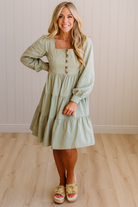 Woman wearing a light green dress with long sleeves in a room with wooden flooring and a white wall.