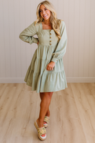 Woman wearing a light green dress with long sleeves in a room with wooden flooring and a white wall.