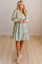Woman wearing a light green dress standing on a wooden floor with a white paneled wall in the background.