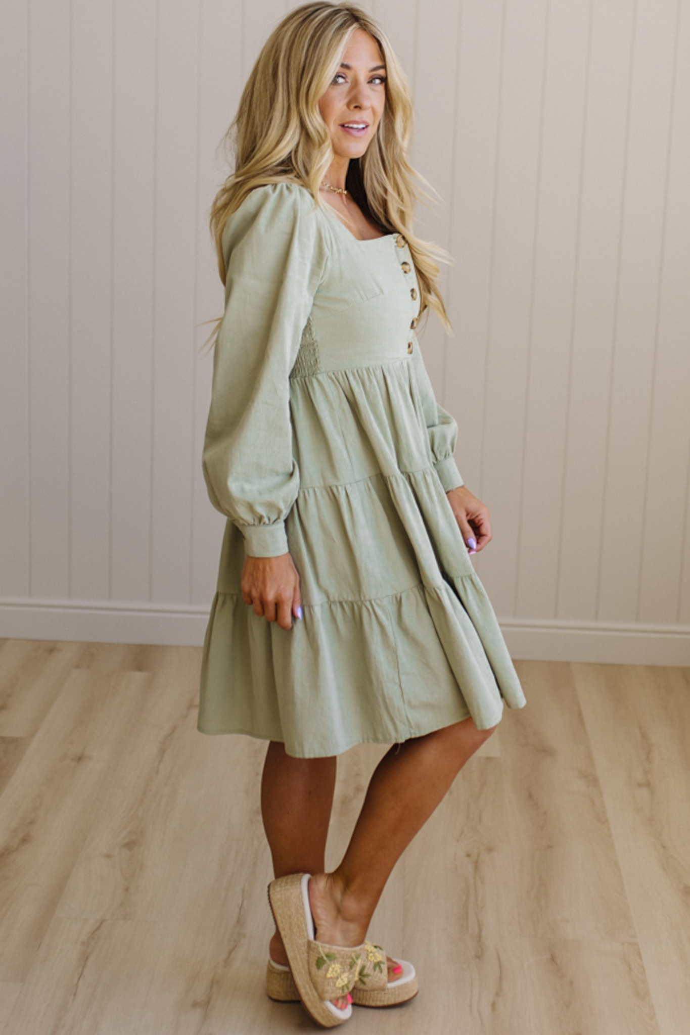 Woman wearing a light green dress with long sleeves in a room with wooden flooring and a white wall.