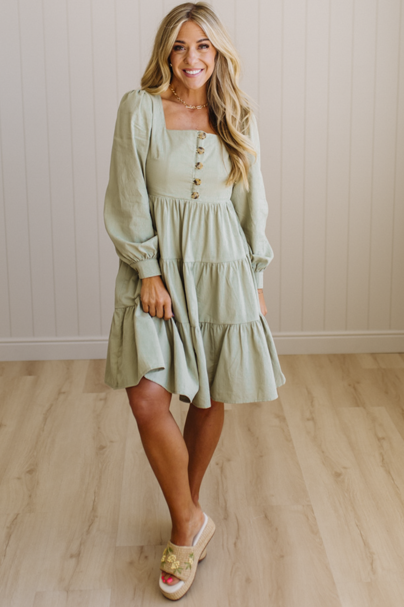 Woman wearing a light green dress with long sleeves and buttons, standing on a wooden floor.