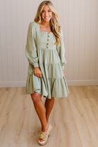 Woman wearing a light green dress with long sleeves and buttons, standing on a wooden floor.