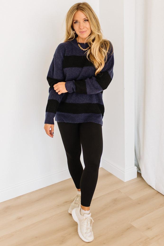 Woman wearing a navy sweater and black leggings with white sneakers against a white wall.