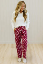 A woman modeling burgundy pants with white polka dots, paired with a white long-sleeve top and white sneakers, standing against a light wood-paneled wall.