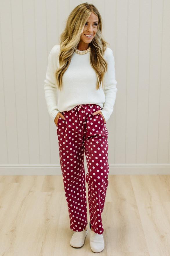 A woman modeling burgundy pants with white polka dots, paired with a white long-sleeve top and white sneakers, standing against a light wood-paneled wall.