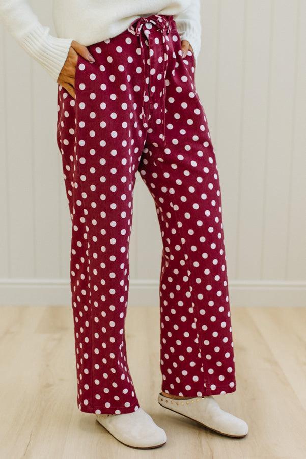 A person wearing burgundy drawstring pants with a white polka dot pattern, styled with a white ribbed sweater and white slip-on shoes in an indoor setting.