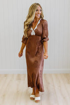 A woman modeling a brown satin maxi dress with a tonal floral pattern, featuring a lace V-neckline and puff sleeves, standing in front of a white paneled wall and light wood floor.