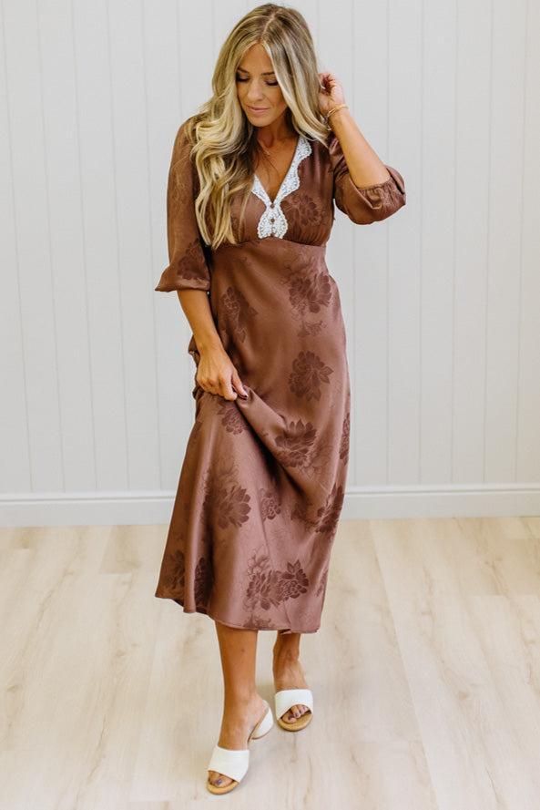 A woman modeling a brown midi-length dress with a subtle floral jacquard pattern, featuring a white lace V-neckline detail, standing on a light wood floor against a white paneled wall.
