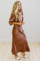 A woman modeling a brown satin-like midi dress with a floral jacquard pattern, featuring a wrap-style collar and three-quarter sleeves, paired with white slide sandals against a white paneled wall and light wood floor.