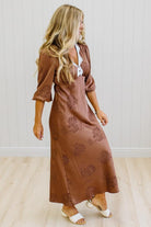 A woman wearing a brown midi dress with a tonal floral pattern, featuring a white lace-up V-neckline detail, standing in a room with light wood floors and white paneled walls.