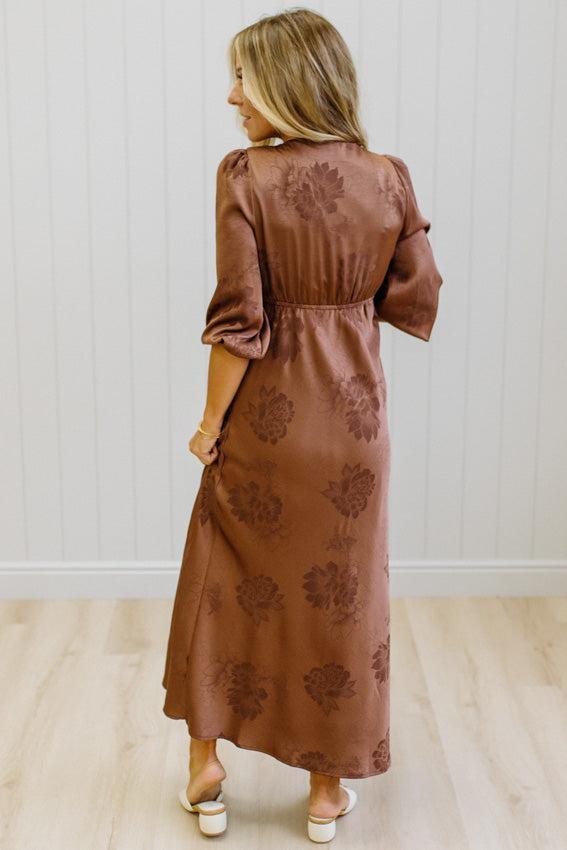 A woman wearing a brown midi dress with a tonal floral jacquard pattern, shown from the back in a bright room with white paneled walls and light wood floors.