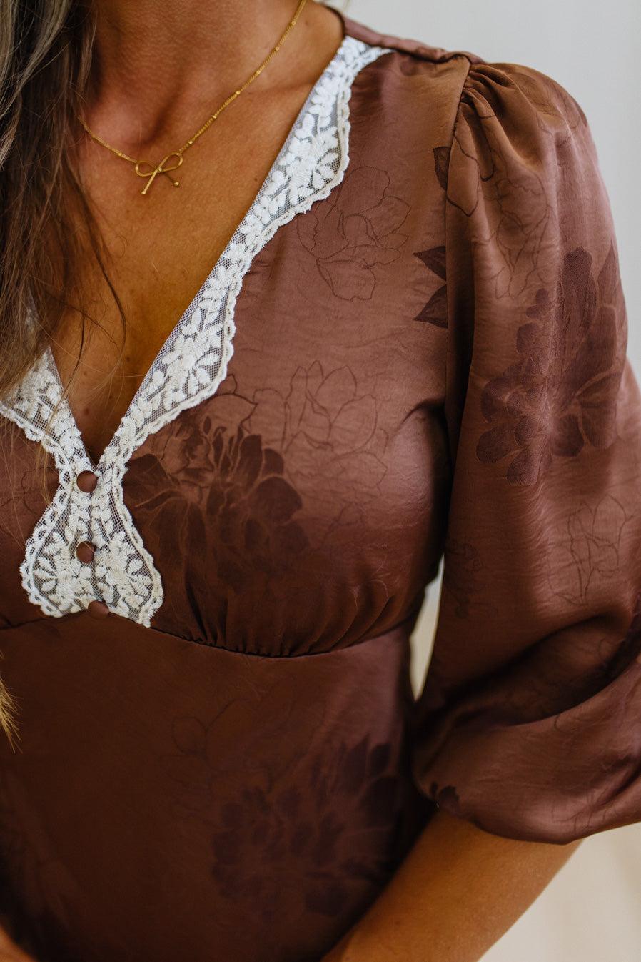 A close-up of a woman wearing a brown satin-like blouse with a floral jacquard pattern, white lace trim along the V-neck and chest, and puff sleeves.