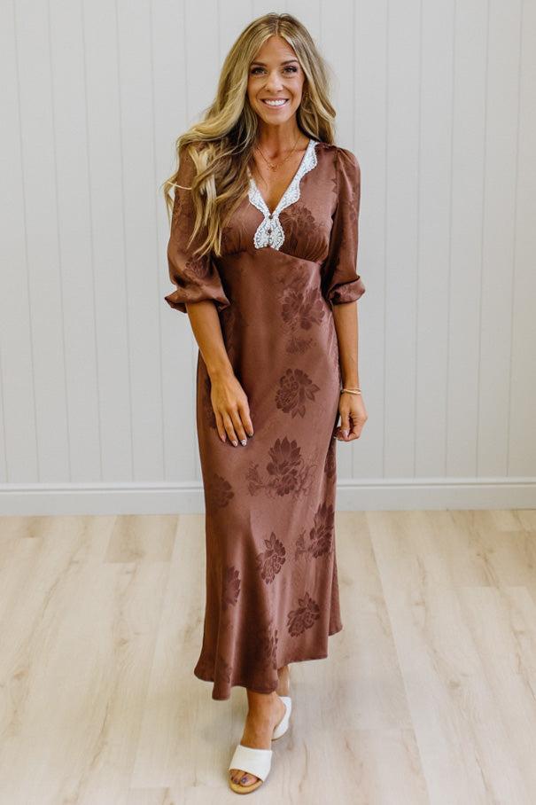 A woman modeling a brown satin maxi dress with a floral jacquard pattern, featuring a lace-trimmed V-neckline and puff sleeves, standing in a room with light wood floors and white paneled walls.