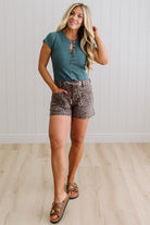 Woman wearing a teal top and patterned shorts standing against a white paneled wall.