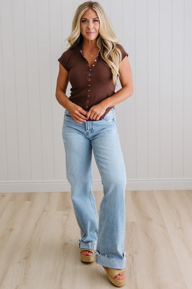 Woman wearing a brown top and light blue jeans standing in a room with a white paneled wall.