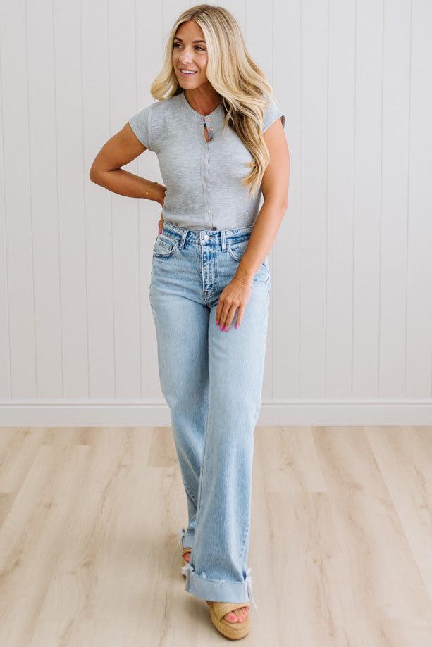 Woman wearing a gray top and light blue jeans standing against a white wall.