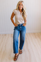 Woman wearing a beige top and blue jeans standing in a room with a white wall and wooden floor.