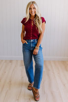 Woman wearing a red top and blue jeans standing against a white paneled wall.