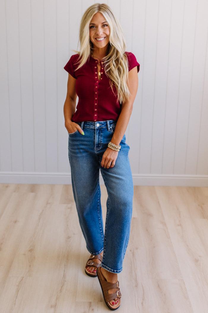 Woman wearing a red top and blue jeans standing against a white paneled wall.