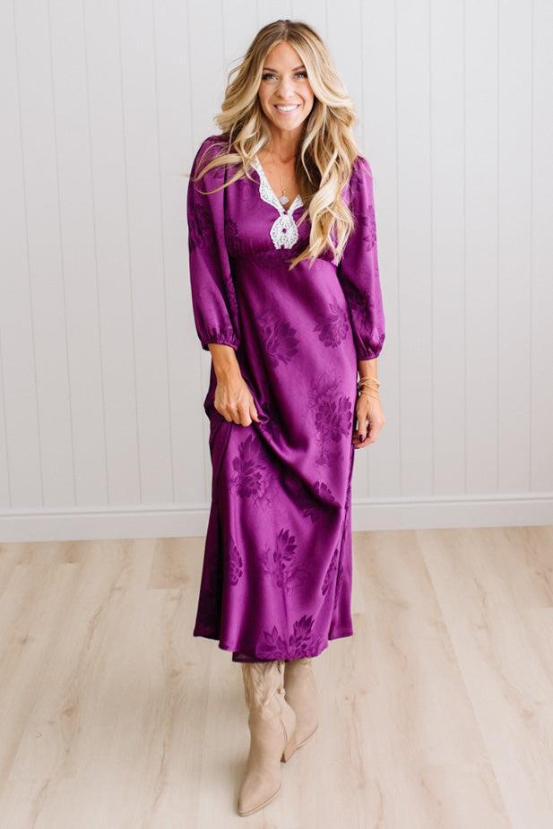 A woman modeling a plum-purple satin-like midi dress with a floral jacquard pattern, featuring a wrap-style collar and three-quarter sleeves, paired with white slide sandals against a white paneled wall and light wood floor.
