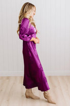 A woman modeling a plum-purple satin-like midi dress with a floral jacquard pattern, featuring a wrap-style collar and three-quarter sleeves, paired with white slide sandals against a white paneled wall and light wood floor.