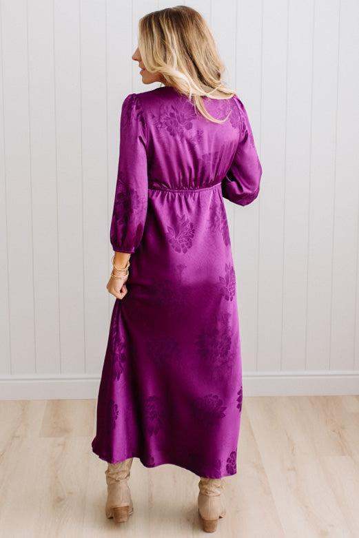 A woman modeling a plum-purple satin-like midi dress with a floral jacquard pattern, featuring a wrap-style collar and three-quarter sleeves, paired with white slide sandals against a white paneled wall and light wood floor.