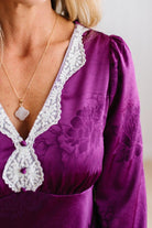 A woman modeling a plum-purple satin-like midi dress with a floral jacquard pattern, featuring a wrap-style collar and three-quarter sleeves, paired with white slide sandals against a white paneled wall and light wood floor.