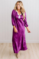 A woman modeling a plum-purple satin-like midi dress with a floral jacquard pattern, featuring a wrap-style collar and three-quarter sleeves, paired with white slide sandals against a white paneled wall and light wood floor.