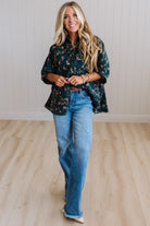A woman stands in a bright room with light wood flooring and white paneled walls, wearing a dark green floral button-up shirt and light blue jeans.
