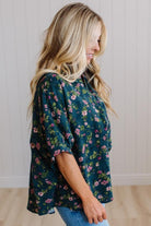 A woman wearing a dark teal green floral blouse with pink and green flowers, paired with blue jeans, standing in a room with white paneled walls and light wood floors.