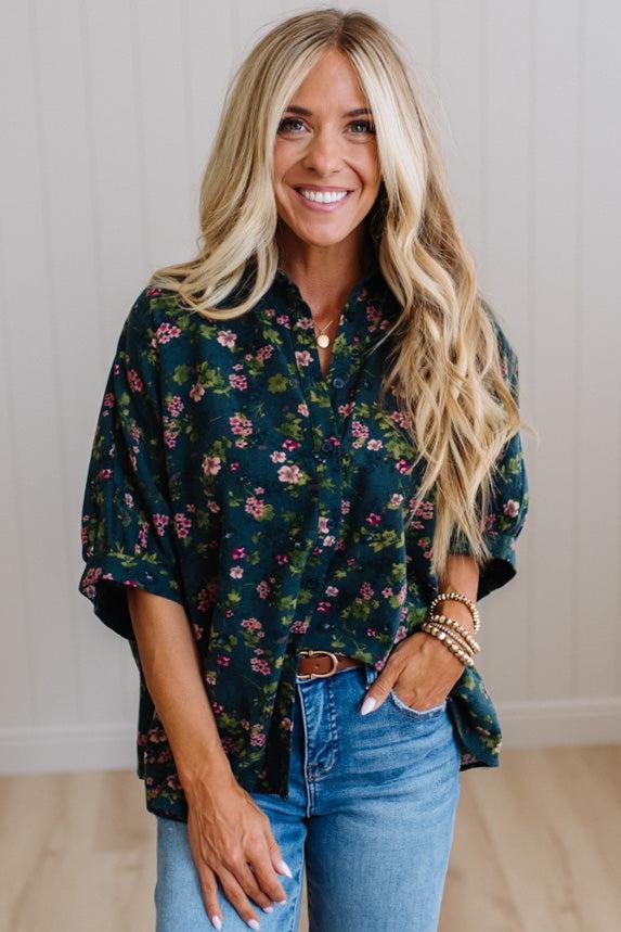 A smiling blonde woman wearing a dark green floral button-up shirt with rolled sleeves and blue jeans, standing against a light paneled wall.