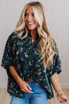 A woman with long blonde hair smiling while wearing a dark green short-sleeve button-up blouse with a pink and green floral pattern, paired with blue jeans.