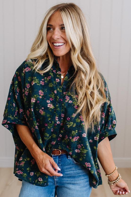 A woman with long blonde hair smiling while wearing a dark green short-sleeve button-up blouse with a pink and green floral pattern, paired with blue jeans.