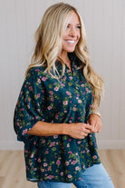 A smiling blonde woman wearing a dark green floral blouse with pink and purple flowers, paired with light blue jeans, standing against a light wood-paneled wall.