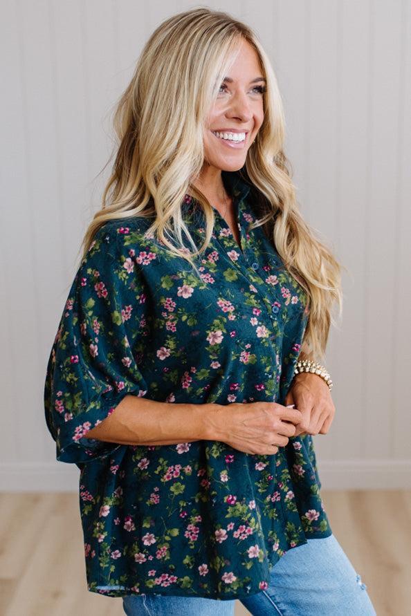 A smiling blonde woman wearing a dark green floral blouse with pink and purple flowers, paired with light blue jeans, standing against a light wood-paneled wall.