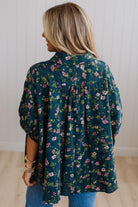 A woman viewed from the back wearing a dark teal-green floral blouse with pink and white flowers, paired with blue jeans, standing against a white paneled wall.