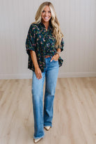 A woman stands in a bright room wearing a dark green floral button-up shirt and light blue wide-leg jeans.